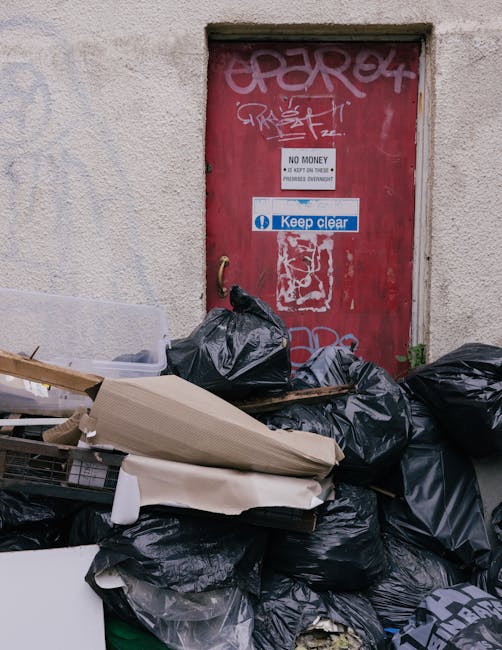 A collection of refuse bags, primarily black, piled up in front of a red door with visible graffiti, signs, and stickers, situated against a textured beige external wall. The refuse bags appear to contain various household waste and are stacked haphazardly, with some leaning or partially overlapping each other. Among the waste, there are discarded cardboard pieces, a piece of white packaging, and a portion of a wire cage or crate peeking out. The red door has several graffiti tags, a small rectangular sign stating 'NO MONEY', and a blue and white sticker reading 'Keep clear'. The scene suggests an outdoor rubbish accumulation area, possibly related to private waste collection, as indicated by the presence of rubbish clearance services like Rubbish Clearance Brompton. The environment is lit with natural daylight, highlighting the textures of the bags, wall, and graffiti-stained door, emphasizing the importance of proper waste disposal and alternative rubbish removal methods.