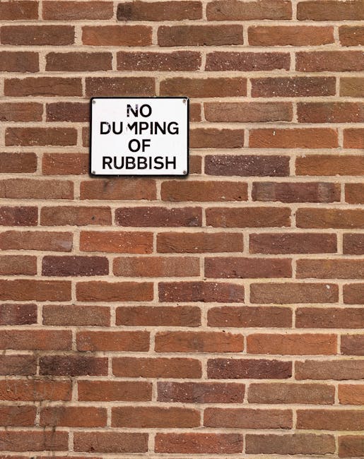A white rectangular sign with black text mounted on a red brick wall, reading 'NO DUMPING OF RUBBISH.' The brick wall features a uniform pattern of reddish-brown bricks with darker mortar joints, and the sign is positioned roughly in the upper left quarter of the image. The surface of the wall has a slightly textured appearance, with some bricks exhibiting variations in tone and finish. The lighting suggests natural daylight, casting a subtle shadow from the sign onto the bricks below. The image emphasizes a clear message against illegal waste disposal in an area that may be used for external rubbish collection or waste management, consistent with services provided by companies like Rubbish Clearance Brompton, and subtly supports the context of maintaining clean, waste-free environments through proper disposal or private clearance services.