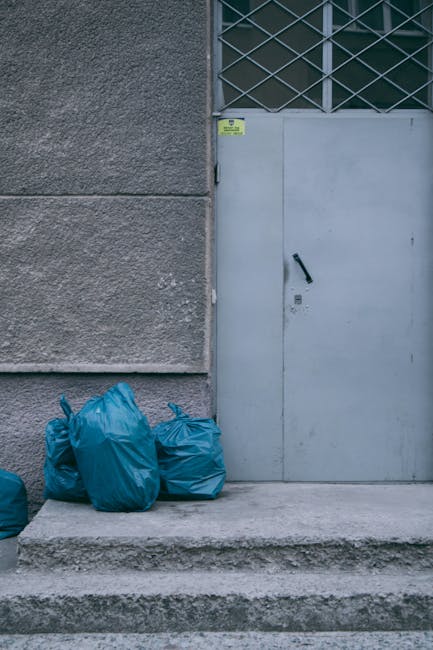 A collection of refuse bags, primarily black, piled up in front of a red door with visible graffiti, signs, and stickers, situated against a textured beige external wall. The refuse bags appear to contain various household waste and are stacked haphazardly, with some leaning or partially overlapping each other. Among the waste, there are discarded cardboard pieces, a piece of white packaging, and a portion of a wire cage or crate peeking out. The red door has several graffiti tags, a small rectangular sign stating 'NO MONEY', and a blue and white sticker reading 'Keep clear'. The scene suggests an outdoor rubbish accumulation area, possibly related to private waste collection, as indicated by the presence of rubbish clearance services like Rubbish Clearance Brompton. The environment is lit with natural daylight, highlighting the textures of the bags, wall, and graffiti-stained door, emphasizing the importance of proper waste disposal and alternative rubbish removal methods.