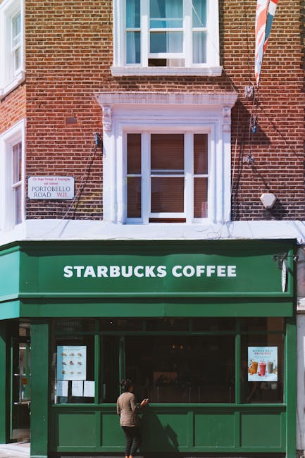 The image depicts the exterior of a building with a brick facade and a white-framed, single-pane window on the upper floor. Below the window, there is a storefront with a green facade and a matching green awning that displays the white text 'STARBUCKS COFFEE'. The storefront has large glass windows and a door, through which the interior of the coffee shop is partially visible. In the foreground, a person dressed in beige pants and a dark top is standing just outside the entrance, looking at their phone. The building is situated on a street corner, indicated by a small street sign on the left side of the image that reads 'PORTOBELLO ROAD, W11'. The scene is outdoors during daytime, with natural light illuminating the brickwork, signage, and sidewalk area. This setting suggests a typical urban location that may be associated with commercial activity, including potential urban rubbish removal or waste management services in the vicinity, reflecting the context of private waste handling around shops and cafes.