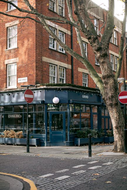 A street scene showing a corner building with a dark blue exterior and large glass windows, which appears to be a café or shop with signage reading 'GRIND' above the entrance. The establishment is situated at the base of a multi-storey, red-brick residential building with white window frames and multiple stories. In the foreground, there are two black bollards and two circular 'no entry' traffic signs attached to metal posts, positioned on either side of the entrance. To the right of the building, a large mature tree with thick, textured bark and extending branches partially obscures the upper part of the structure. The tree's trunk and branches are visible, with some branches stretching over the street. The pavement and street are paved with cobblestones, and the street corner features a small section of double yellow lines indicating parking restrictions. The scene is illuminated by natural daylight with slight shadows, creating a calm urban atmosphere. Rubbish clearance services by Rubbish Clearance Brompton may be relevant in the context of maintaining clean, clutter-free commercial and residential frontages in the area, supporting independent waste collection or on-site clearance options.