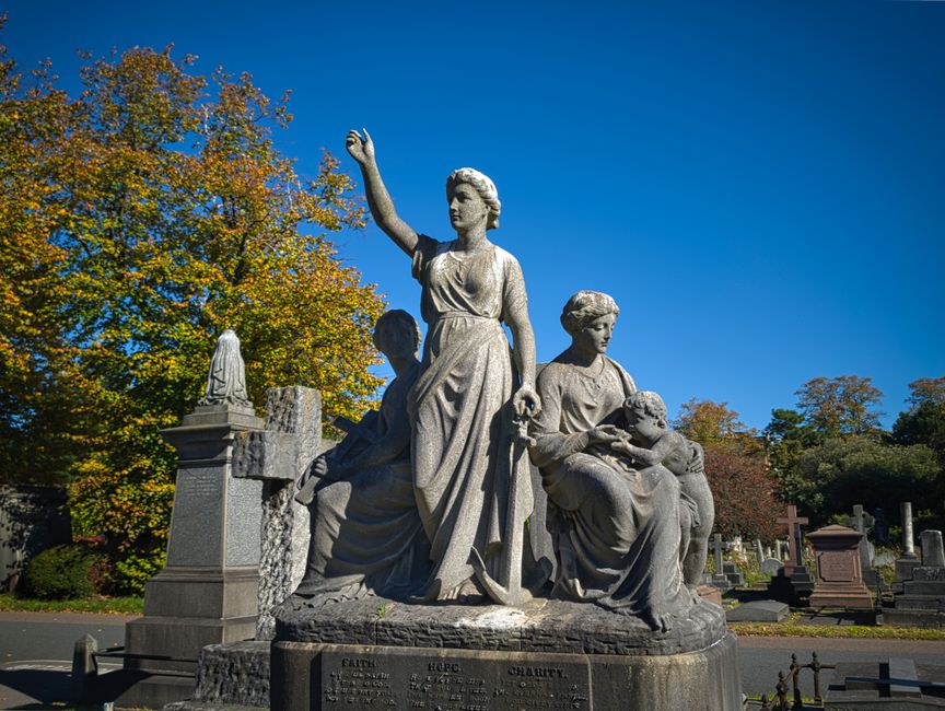 A large light grey stone monument featuring three intricately carved female figures set in a cemetery. The central figure stands upright with her right arm raised and hand open, wearing a flowing dress with detailed folds. To her left, a seated woman is depicted holding a child, gazing downward, while another woman, also seated, appears to be resting her arm on the child's head. The stone monument has a textured surface with visible weathering, and the base includes engraved inscriptions. Surrounding the monument are other tombstones and grave markers of varying heights and materials, some with crosses and statues. In the background, there is a lush arrangement of trees with green and autumn-colored leaves under a bright blue sky, casting natural daylight on the scene. The setting suggests a peaceful cemetery environment, with the monument being a prominent focal point, indicative of memorial or tribute, typical in burial grounds. The scene aligns with the context of alternative rubbish disposal in cemetery settings or private heritage site maintenance, as managed by local waste removal services like Rubbish Clearance Brompton.