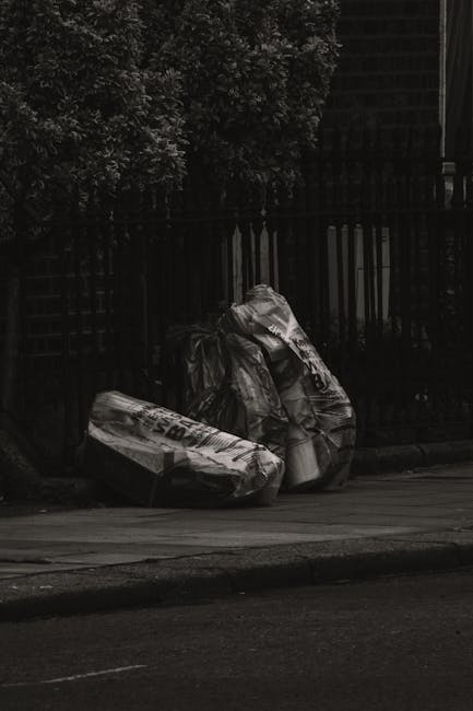 Two large black plastic rubbish bags filled with waste are placed on the pavement beside a black metal fence, with one bag leaning slightly against the other. The bags appear to be tightly knotted at the top and are situated in a residential or urban outdoor setting. Behind the fence, dense foliage and trees are visible, suggesting a garden or private property boundary. The scene is captured in natural daylight, with shadows cast on the ground. The presence of the rubbish bags indicates a collection or disposal process, which could be related to alternative waste handling by private rubbish removal services such as Rubbish Clearance Brompton, rather than council collection. The bags' plastic finish and the partially visible markings or text on them hint at their disposable nature, suitable for domestic or small-scale commercial rubbish removal as part of on-site clearance efforts.