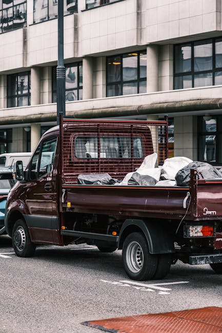 A small, dark red flatbed truck parked on an urban street in front of a modern office building with large glass windows and a white stone facade. The truck's cargo bed is loaded with various waste items, including black and white plastic bags, some of which are partially open, revealing discarded materials inside. The truck has a protective metal mesh on the cab's rear window and side panels, indicating it is used for rubbish or waste collection. The scene suggests an environment where private waste removal services, such as those by Rubbish Clearance Brompton, are employed for on-site rubbish clearance or alternative disposal solutions outside of standard municipal collection. The weather appears overcast, with neutral ambient lighting highlighting the details of the vehicle and the surrounding urban setting. The street surface is grey tarmac with visible road markings, and nearby parked vehicles indicate a typical city environment for waste removal operations focused on commercial or residential rubbish clearance.