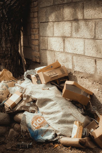 Two large black plastic rubbish bags filled with waste are placed on the pavement beside a black metal fence, with one bag leaning slightly against the other. The bags appear to be tightly knotted at the top and are situated in a residential or urban outdoor setting. Behind the fence, dense foliage and trees are visible, suggesting a garden or private property boundary. The scene is captured in natural daylight, with shadows cast on the ground. The presence of the rubbish bags indicates a collection or disposal process, which could be related to alternative waste handling by private rubbish removal services such as Rubbish Clearance Brompton, rather than council collection. The bags' plastic finish and the partially visible markings or text on them hint at their disposable nature, suitable for domestic or small-scale commercial rubbish removal as part of on-site clearance efforts.